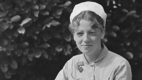 A black and white photo taken of Ann Remmers as a young midwife. She is wearing hospital scrubs and a white cap.  Her curly hair is pulled back and tucked underneath her cap, with a few pieces pulled out at the front.