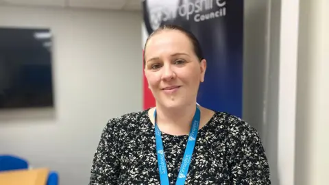 A woman with brown hair tied back, wearing a black top with white flowers on it. She has a blue Shropshire Council lanyard around her neck