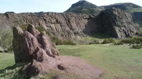 A large jaggy piece of red rock sticks up from the ground. there is green grass around it and cliffs behind it. In the distance is Arthur's Seat.