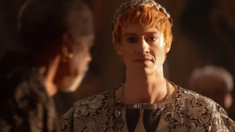 Paramount Pictures A man with red hair wearing a gold and black cloak and a gold crown around his red hair.