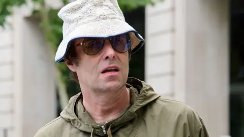 Liam Gallagher, wearing a white lace-patterned bucket hat, sunglasses and a green hooded top, stands in front of a white bricked hotel