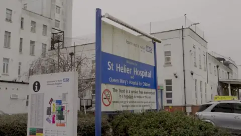 BBC The St Helier Hospital sign outside the hospital, looking dirty, with the wording 'St Helier Hospital and Queen Mary's Hospital for Children'. The large board is blue and white. 