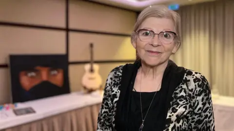 Andrea is wearing a black and white animal-print top with a black scarf-like neckline stands in a conference room. Behind them is a table with a blue bucket, papers, and a guitar propped against the wall. The room has beige walls and a ceiling with recessed lighting and purple accents.