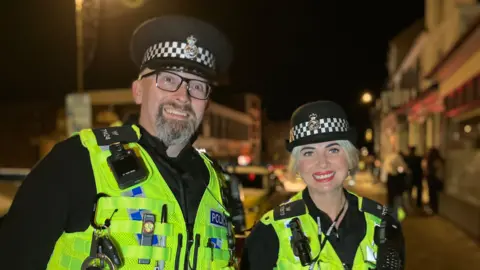 A police officer with a grey beard and bright vest stood next to a female police officer with a police vest.