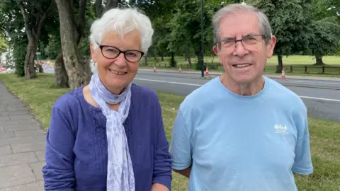 Sofía Luis-Hobbs/BBC A woman wears glasses as she smiles at the camera. She has a purple top on and a white and purple sheer scarf on. To her right is a man with glasses wearing a light blue t-shirt. They are both on a footpath surrounded by greenery on a bright day.