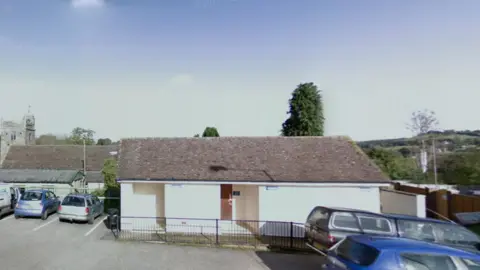 Google street view image of the public toilets in Newton Poppleford. The one-storey block is painted a white colour. Parked cars are to the front and side of the toilet block. 