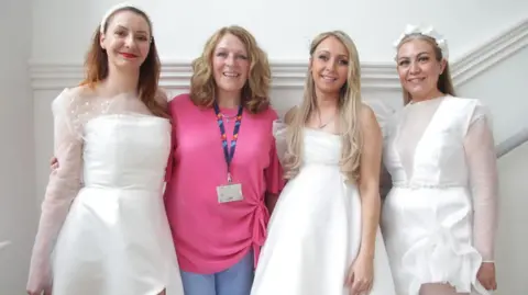 Three woman (models called Ilona Chmiela, Reanna McGuire and Mandy Gibson) in white bridal dresses stand arm in arm. Another woman, second from the left, is casually dressed in a pink top and blue trousers. They are standing in what looks like a stairway with a white wall in the background.