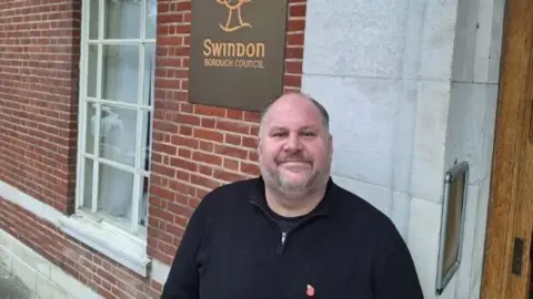 Swindon Borough Council Jim Robbins stands outside the Swindon Borough Council office. He has short grey hair and stubble and wears a black half-zip jumper.