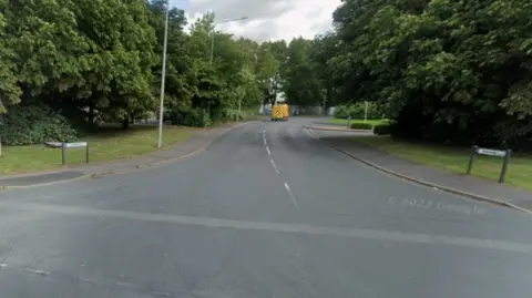 The entrance to Roman Way industrial estate with trees either side of the road. A van with yellow and red hi-vis markings is driving away from the camera.