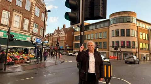 A woman with white hair, wearing a white top and a black jacket and trousers stands in front of a traffic light in the middle of a road junction with tall buildings on all sides. She is pointing up at the traffic light with her right hand and is looking in to the camera