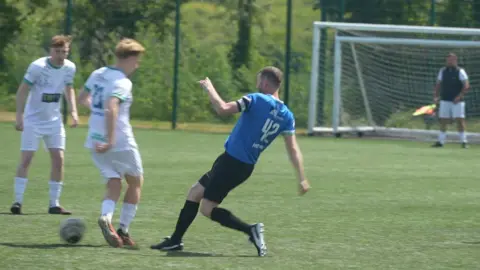 Connor Bennett/BBC Two players in white football kit, with a player in blue making a lunge for the ball. A goalkeeper in black and white stands in goal in the background