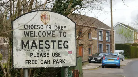  "Welcome to Maesteg, please use free car park". Behind the sign is a row of terraced houses and some parked up cars. 