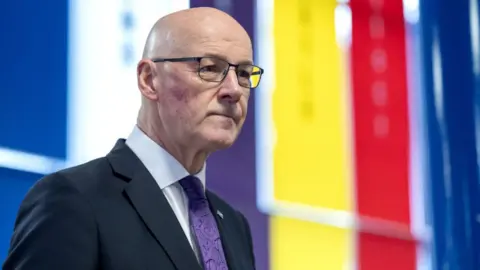 A bald man with glasses stands in front of a striped multi-coloured background. He is wearing a dark suit, white shirt and purple tie. 