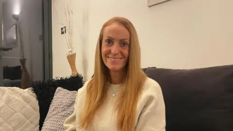 The image shows a middle-aged woman with long blonde straight hair and a white jumper, sitting on her sofa and smiling at the camera.