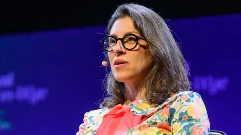 Sam Hardwick/Hay Festival A close up image of a woman with dark hair, wearing a multicoloured floral dress and glasses. 
