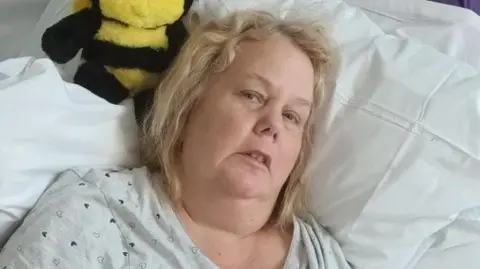 Family handout Donna lies propped up in a hospital bed at Hull Royal Infirmary. She has should length blonde hair and wears a light-coloured hospital. There is a small black-and-yellow soft toy, shaped like a bee, resting on the pillow beside her head.