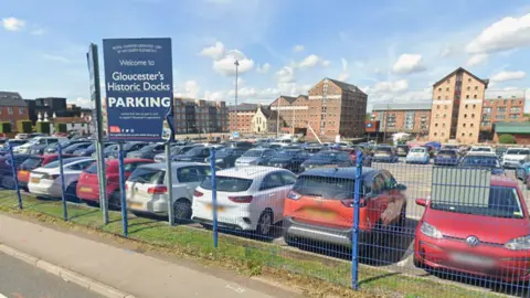 Google A screenshot from Google street view showing the car park at Soutgate Moorings. It is a large space full of cars, surrounded by blue metal fencing and tall residential flat buildings by the water.