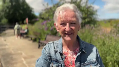An older lady with short white hair, a denim jacket and pink t-shirt. There is shrubbery and park benches in the background.