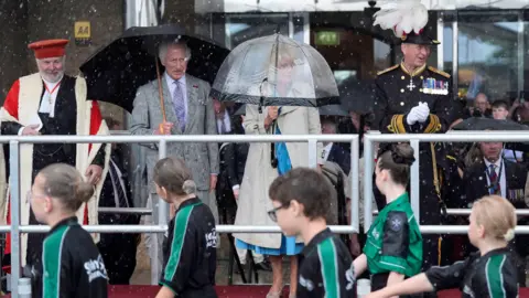 PA Media King Charles and Queen Camilla hold umbrellas as a troop of beaver scouts parade past.