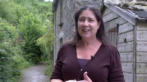 The image shows Emma Eveleigh standing outside the old granite building of Haye Mill with a large area of green trees to her left. She is smiling, wearing a brown cardigan and purple top underneath it.