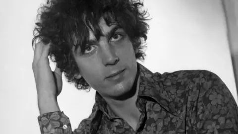 A black and white image of Syd Barrett. He had curling dark hair cut in a fringe and is wearing a floral shirt. His right hand is resting in his hair.