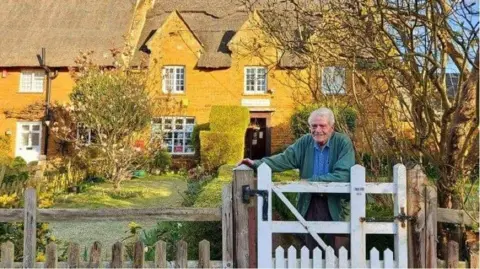 Matt Masters Dave Masters in a green cardigan and blue shirt at a white wooden gate in front of a stone cottage.