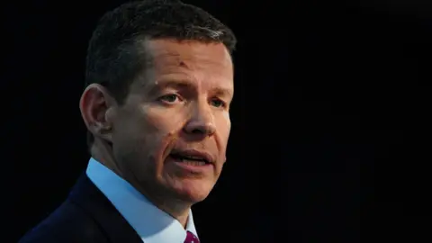 Rhun ap Iorwerth looking to his right hand side, wearing a blue shirt, red tie and dark suit jacket. It is a head and shoulders picture with the background in darkness.