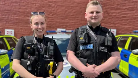 Anna and Callum stand smiling at the camera in the police station car park next to three police cars. They wear black police uniform including the black vest which carries equipment such as walkie talkies and wallets. They both have their clasped in front of their waist. Anna has blonde hair in a ponytail and has sunglasses on her head, while Callum has short fair hair. 