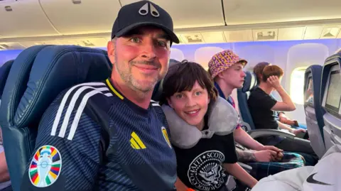 a man wearing a cap and Scotland strip smiling, next to him is a young boy with long brown hair. They are on a plane.