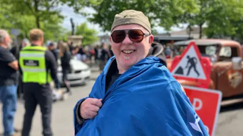 A man with a green cap, sunglasses and wrapped in a large blue cover, smiles. Behind him is a crowd of people with the statue of Lemmy just visible in the distance.