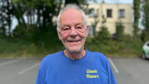 A man with grey whispy hair looking at the camera. He's wearing a t shirt that is blue and has the words Classic Flowers embroidered on it.