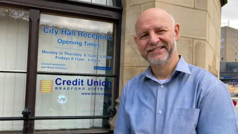 BBC A bald man with a white beard and wearing a blue shirt smiles outside the window where details of Bradford District Credit Union appear on a transparent screen.