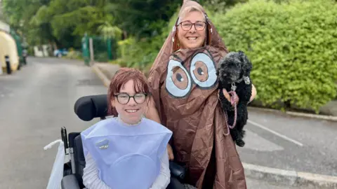 Carmela Chillory-Watson is sittin in a wheelchair dressed as a toilet. She has dark hair which is tied back and is wearing glasses. Her mother, Lucy, is to the left of the image dressed in an emoji poo outfit. She is standing up, holding a black dog under her left arm and is also wearing glasses. The pair are smiling at the camera.