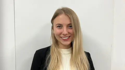 Matt Knight/BBC A head and shoulders picture of Anna Sahan standing in front of a white wall. She has long blonde hair and is smiling.