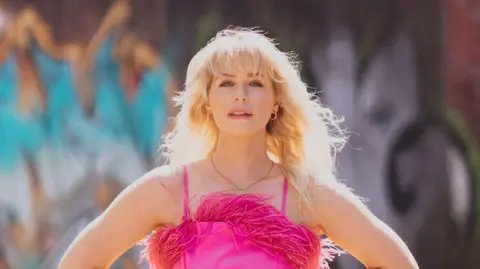 Rhianon Hol Jess Davies wears a neon pink jumpsuit, with feather fringe detailing. She's wearing a gold necklace and hoop earrings, with her wavy blonde hair swept in the wind.
