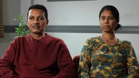 A man with short black hair and wearing a dark red jumper is sat next to a woman with dark hair, tied back, and wearing a gold necklace and a gold and green dress.