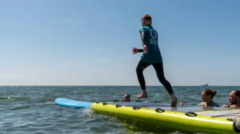 A male is about to jump off a raft and onto a surfboard.