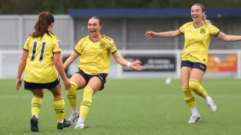Three female footballers all wearing a yellow and dark navy blur football kit seemingly celebrating a goal together on a football pitch.