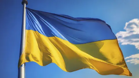 Getty Images A photo of a Ukrainian flag, with a blue upper half and a yellow bottom half flying on a metal flagpole with a blue sky in the background