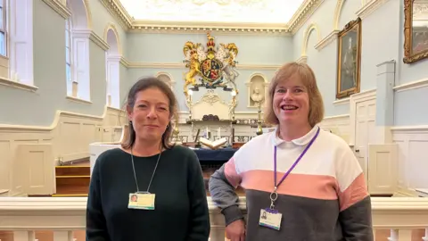 A head and shoulder shot of two women in an ornate old style courtroom with pale blue walls and white wooden panelling. There is a large coat of arms on the back wall showing a golden lion and a white unicorn. The two women have shoulder length hair, one blonde, one brown. One is wearing a dark blue jumper, the other a striped blue, pink and cream jumper. They both have lanyards around their necks.