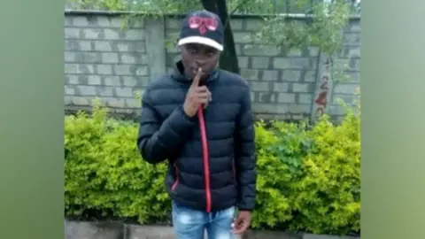 Boniface Kariuki in a black jacket and cap, holding his finger to his lips