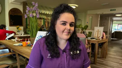 Cathy Killick/BBC A woman with curly black hair smiles at the camera. She wears a purple uniform. Women are sat at tables behind her as they work on canvases.