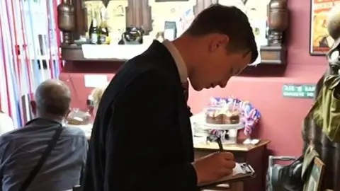 Jodie Halford/BBC Lincoln takes a customer's order in the tearoom. He writes on a note pad.