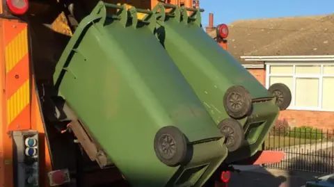 Two green bins being emptied into a lorry. A house and gate can be seen in the background behind the lorry.