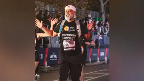 Javad Malik Javad finishes a marthon and waves his hands in joy. He wears a black tshirt with Ramadan Runners on the front, and also the words "World Central Kitchen". He has a white baseball cap on backwards and has a grey beard and moustache. It is quite dark and it looks like the New York marathon