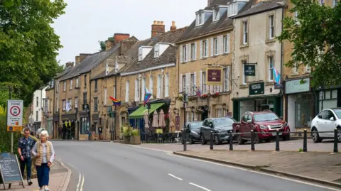 A road in the centre of Chipping Norton. To the right is a row of cafes, pubs and businesses. To the left, two people can be seen walking on the path. 