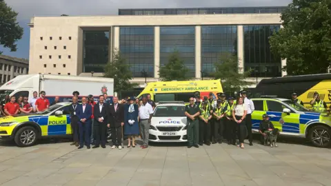 A group of people from various organisations - including police, ambulance, and fire, gather in uniform outside a large, modern building in a city centre square.