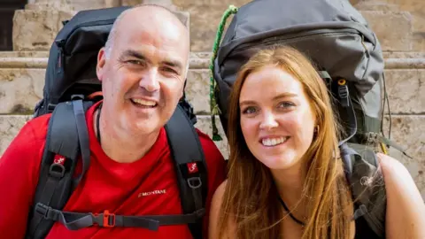 two people looking at the camera - they are a bald man and a red-headed woman. Both are wearing big rucksacks