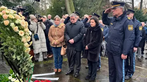 A police officer salutes and other people look on after a wreath of flowers is laid on a rainy day in a park in Bavaria where a toddler and a man were fatally attacked.