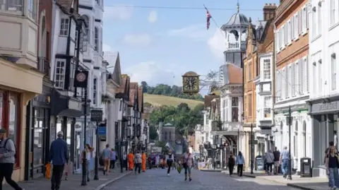 An view down a small high street lined with historic buildings. In the background are green hills and trees